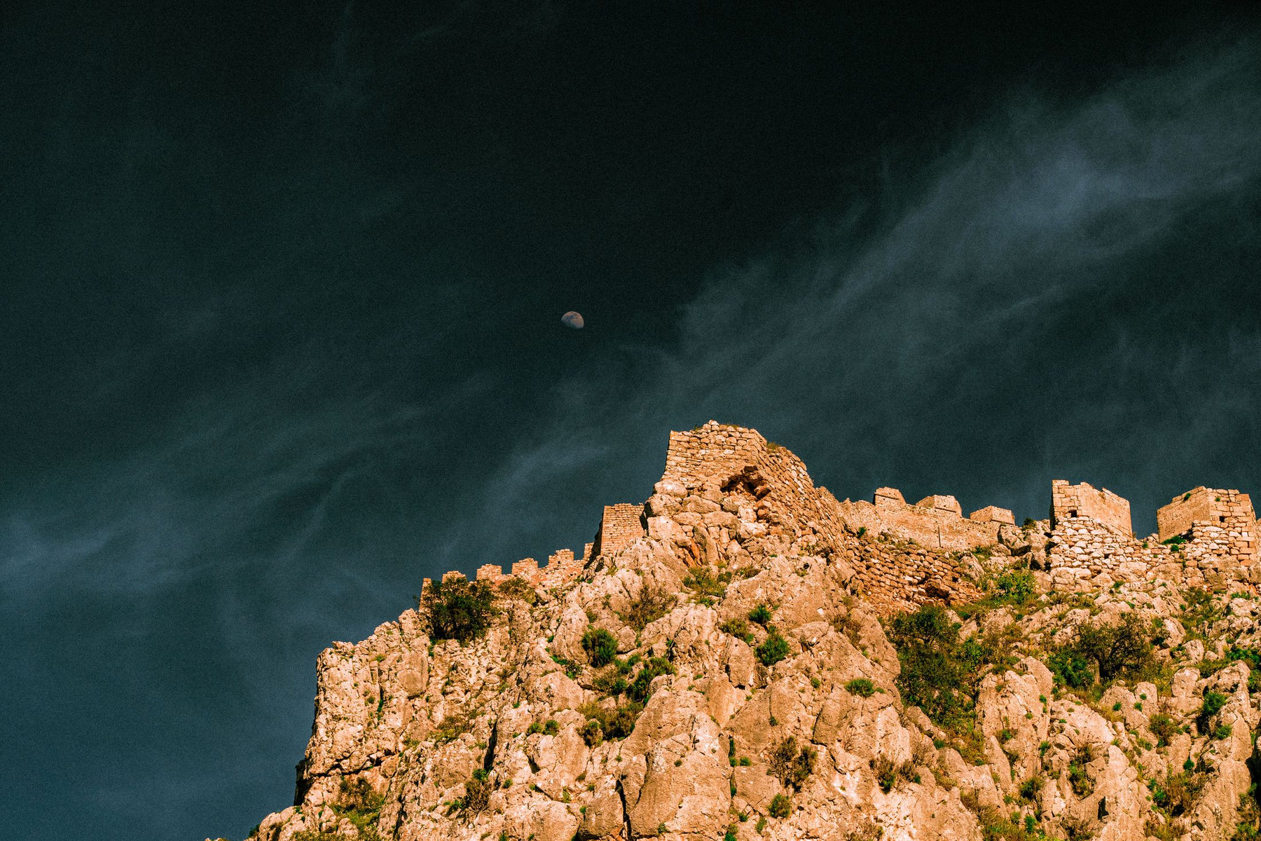 Fortress walls along a ridge under a dark sky, the moon just above the stone